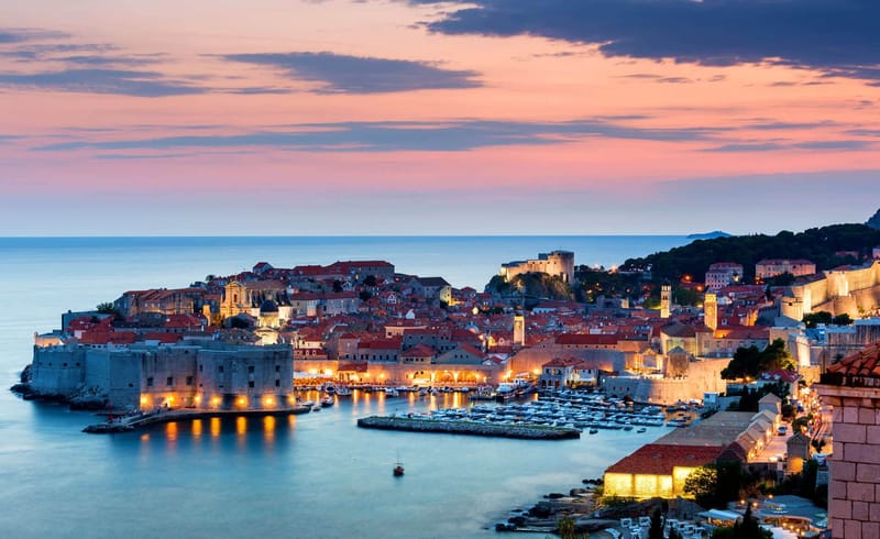 Walled medieval town illuminated at dusk with fortified harbour, moored boats, and pink sunset sky reflecting on calm water