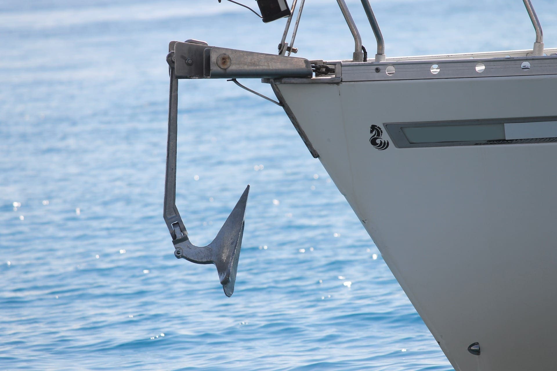 Yacht anchor hanging from the bow roller above clear blue water, showing the shank, crown, and flukes — the components that dig into the seabed to hold the boat in position