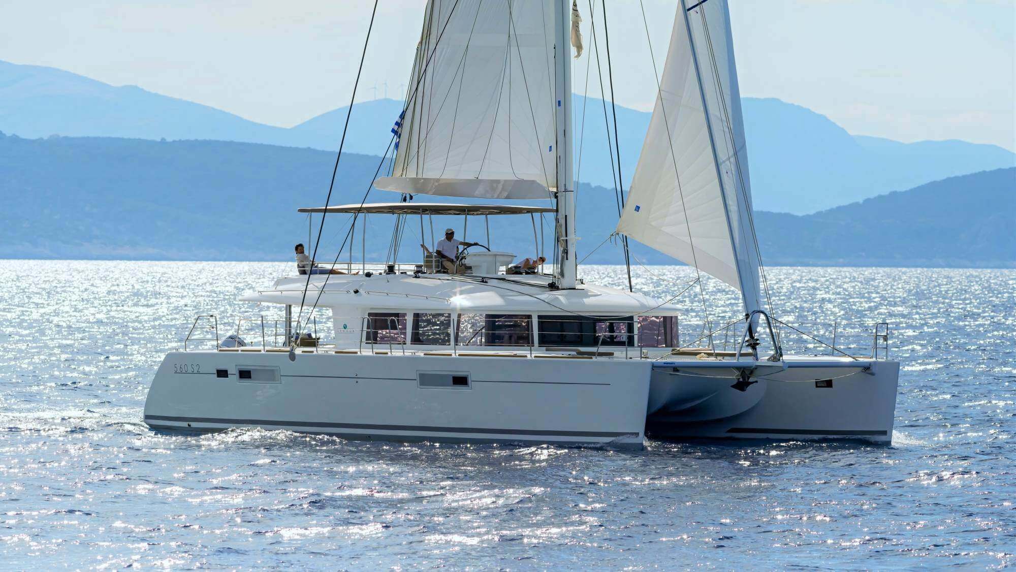 Lagoon 560 catamaran under sail with large overlapping genoa deployed on the forestay, crew visible on deck against mountainous coastline — a genoa is the large headsail that extends past the mast, adding sail area and power in light to moderate winds