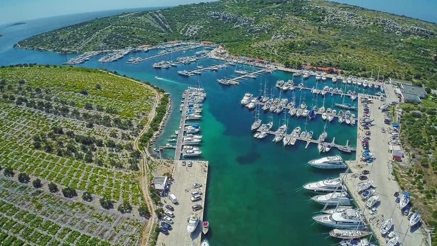 Aerial view of a Mediterranean marina showing rows of sailing yachts berthed stern-to on floating pontoons in turquoise water — a marina provides permanent berths, shore power, and water for visiting boats